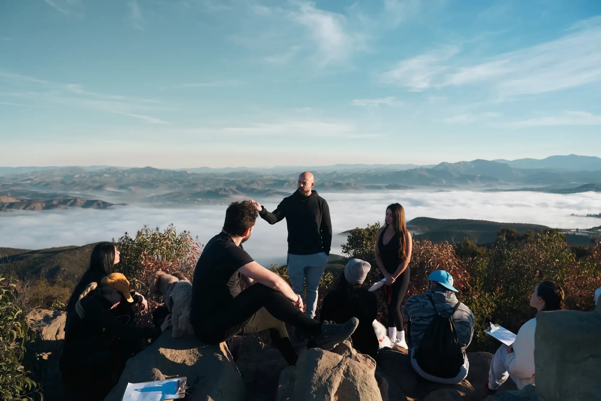 Garin Heslop standing on a mountaintop above a sea of clouds, talking with a small group seated on the rocks around him