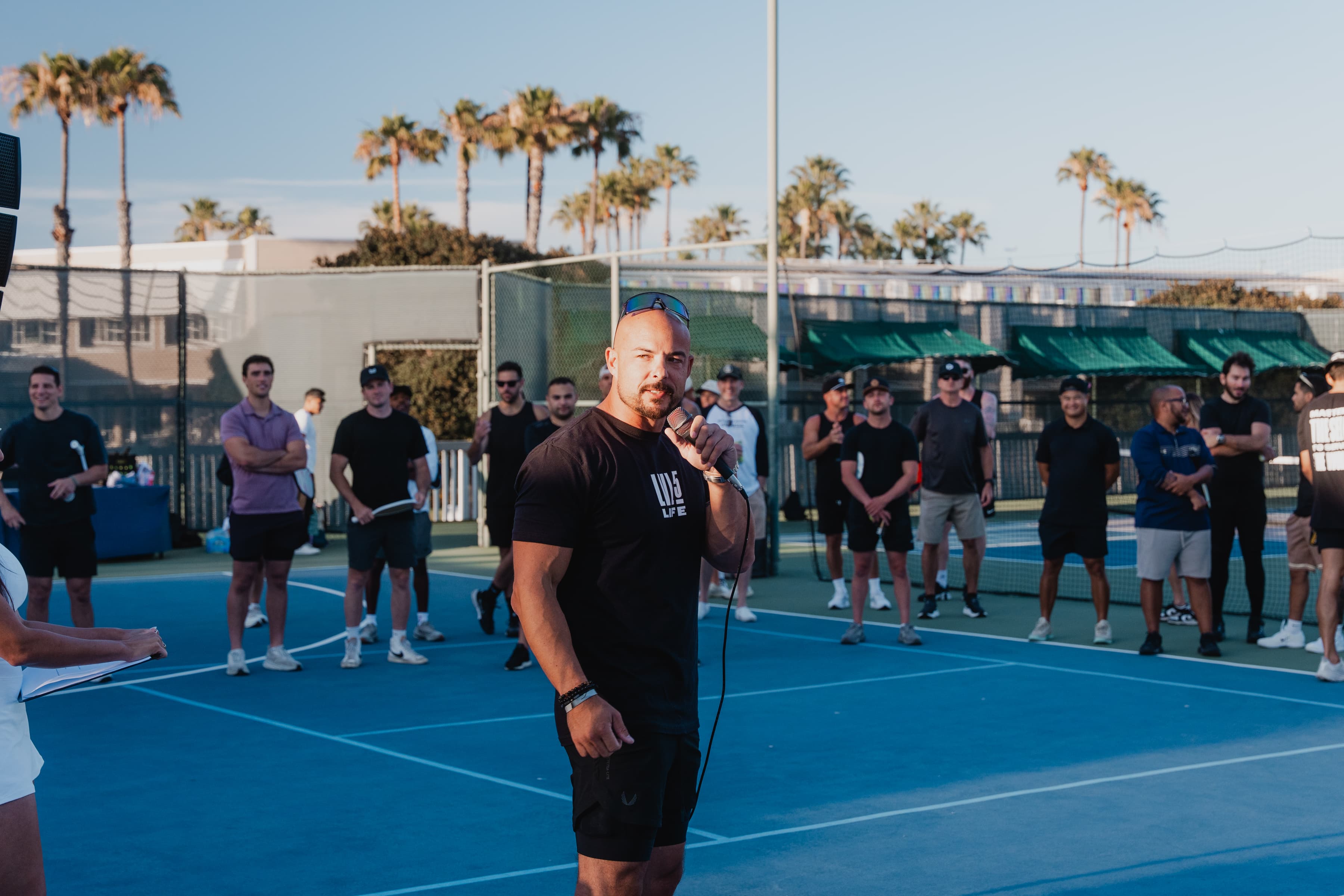 Garin Heslop speaking into a microphone to a group gathered on outdoor tennis courts lined with palm trees