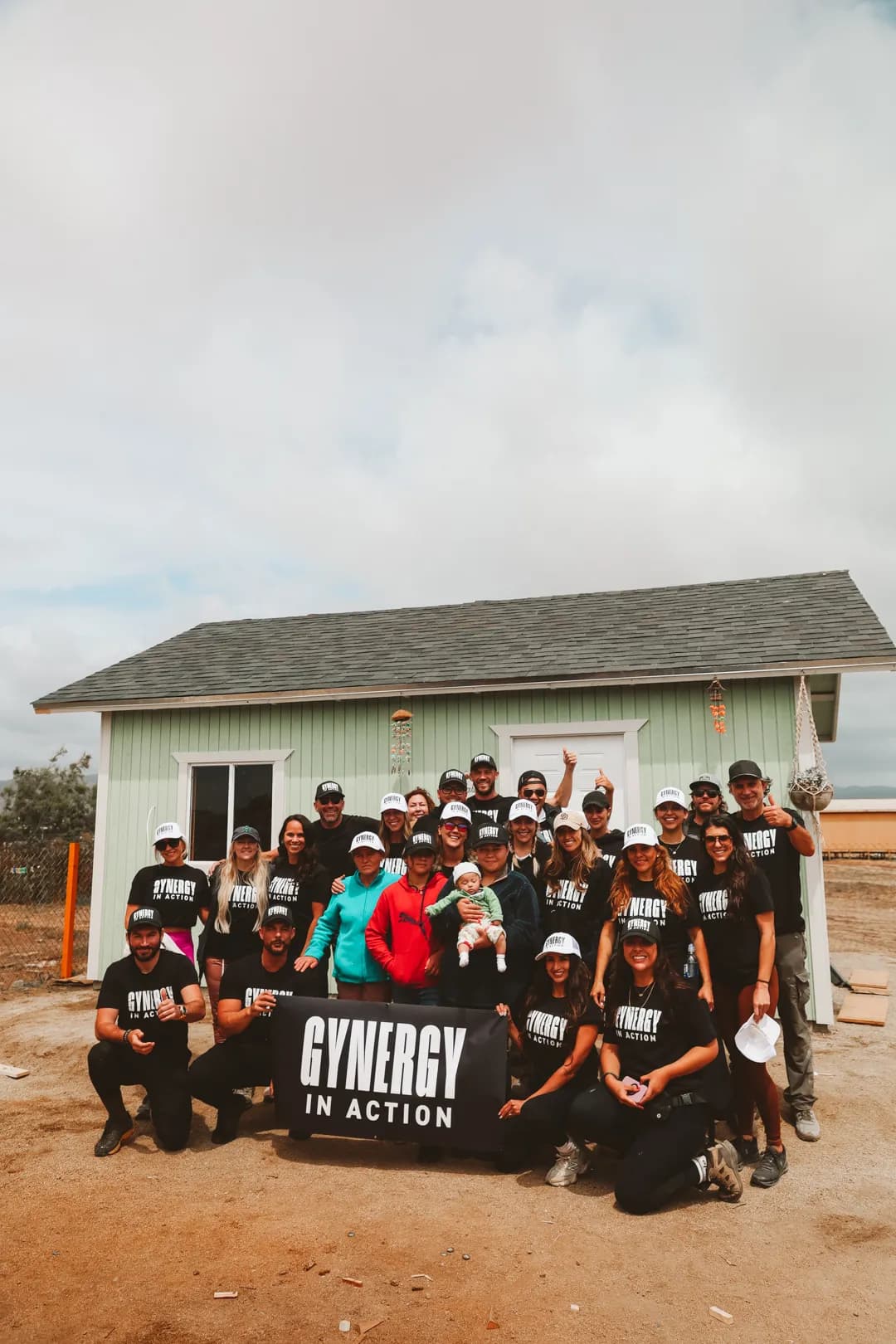 The build crew in black GYNERGY In Action shirts with the family, holding their baby, in front of the light-green home they built, behind a GYNERGY In Action banner